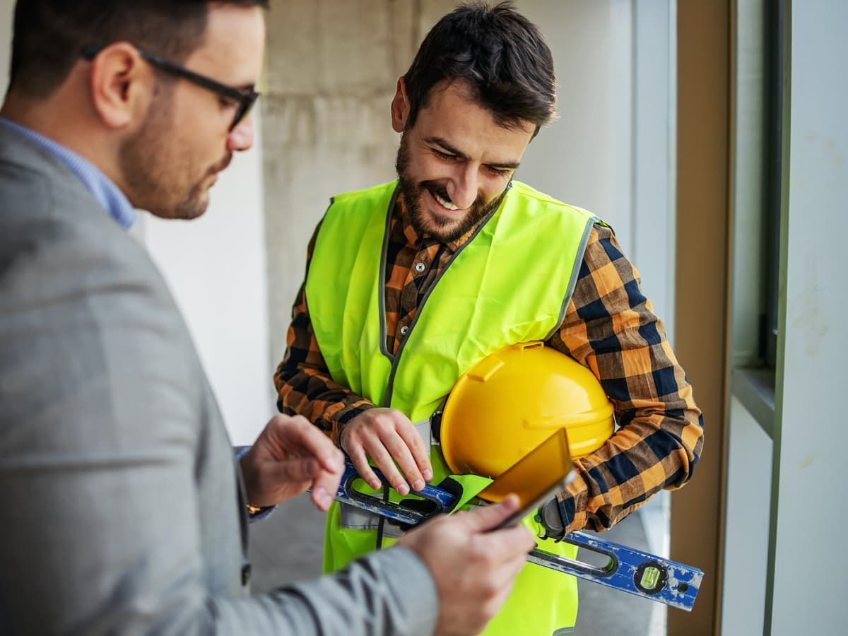 Manager and construction worker with hard hat and vest examining a tablet.