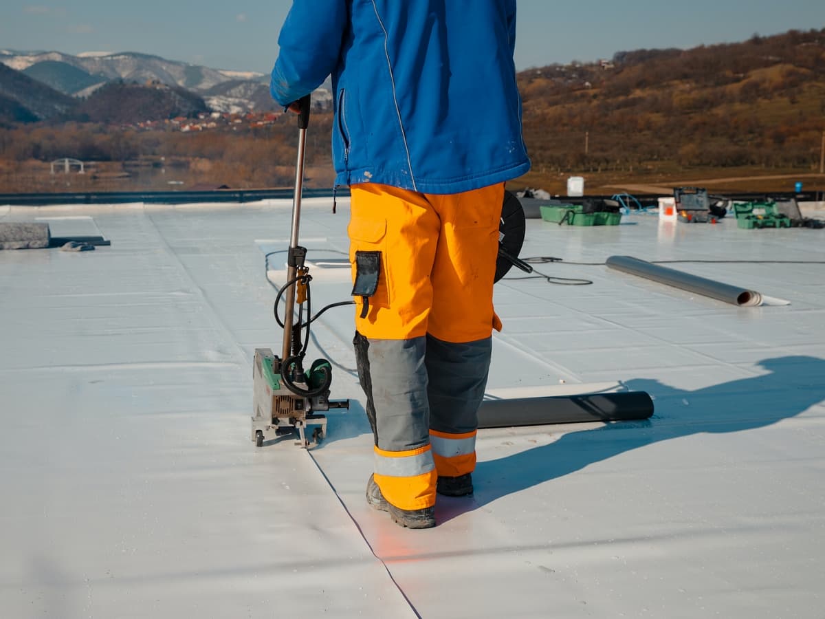 A man in orange pants and a blue shirt stands on a rooftop, working with installation tools.