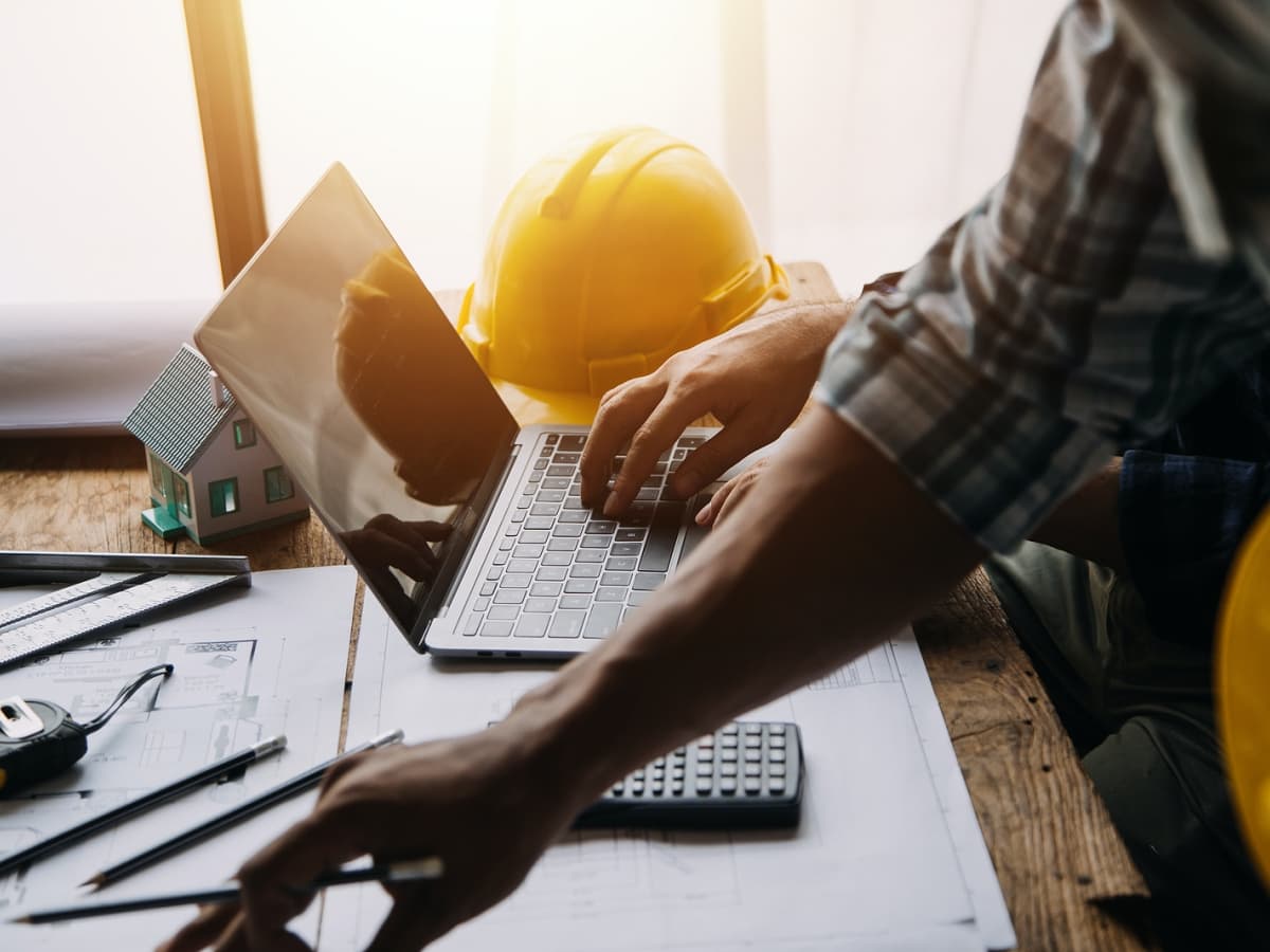 A man in a hard hat and yellow helmet focused on his laptop while working on a construction site.