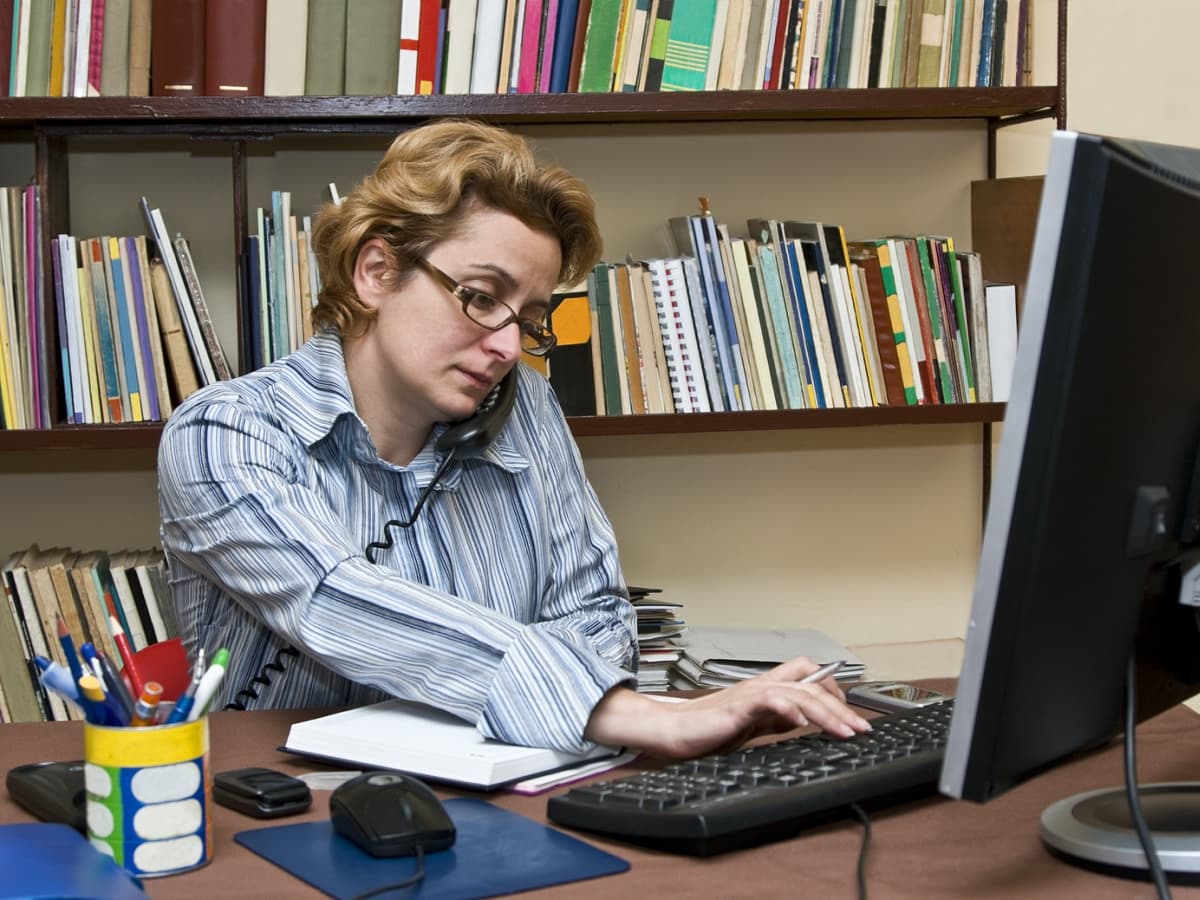 A professional woman wearing glasses is seated at a desk, focused on her computer screen.