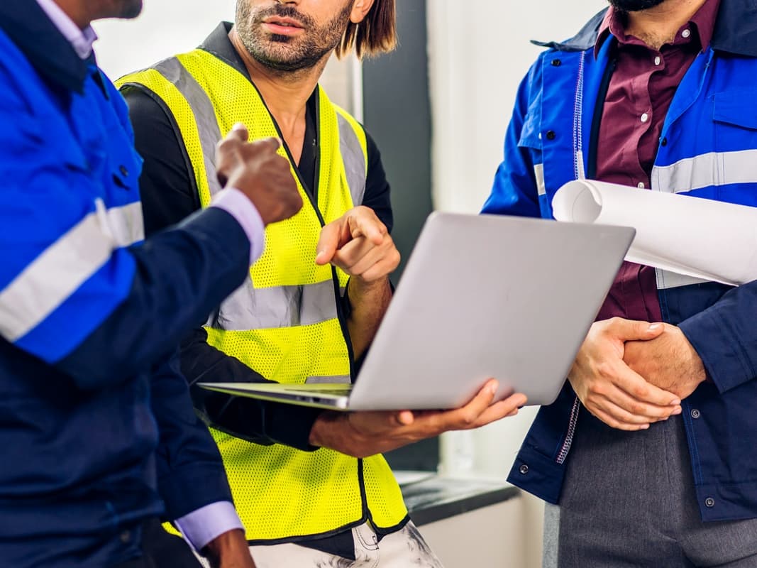 Three men in blue vests and hard hats gathered around a laptop, engaged in focused discussion.