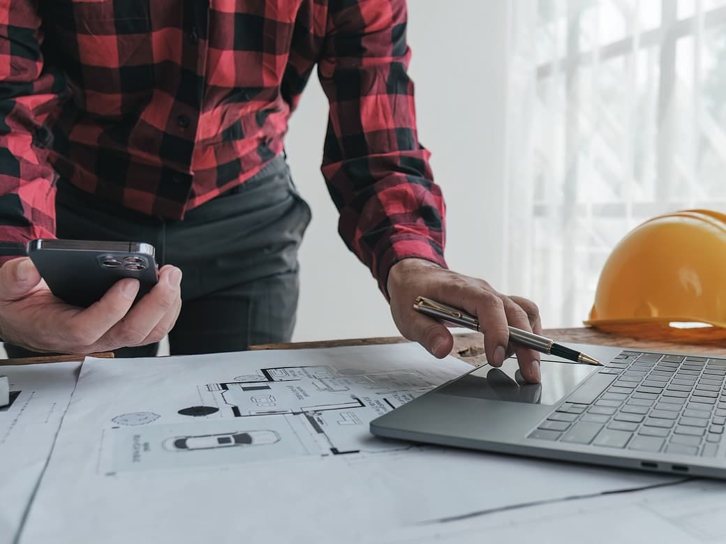 A man in a hard hat and plaid shirt multitasks with a laptop and cell phone.