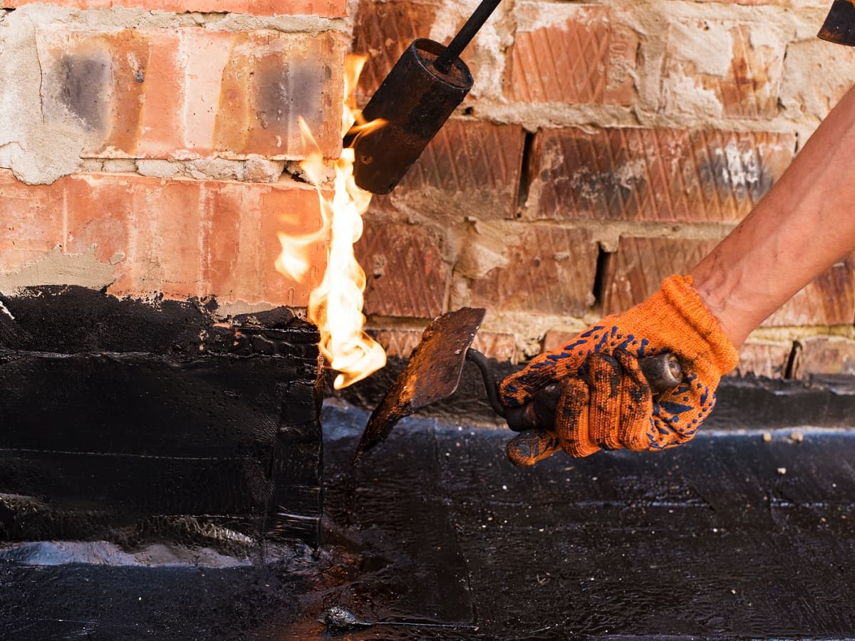 Roofer with orange gloves using flaming tool to apply tar to bricks.