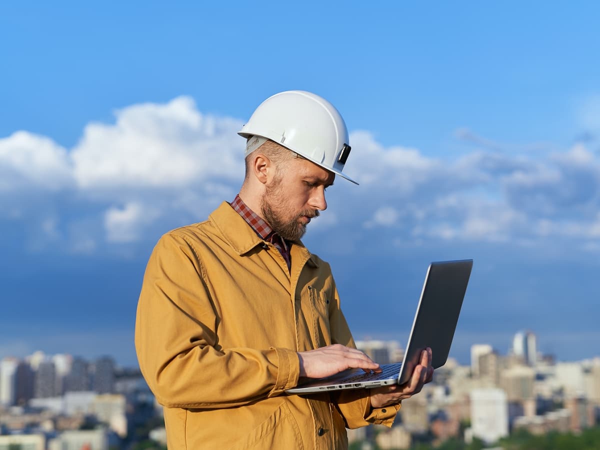 A man in a hard hat and yellow jacket using a laptop to perform tasks at a construction site.