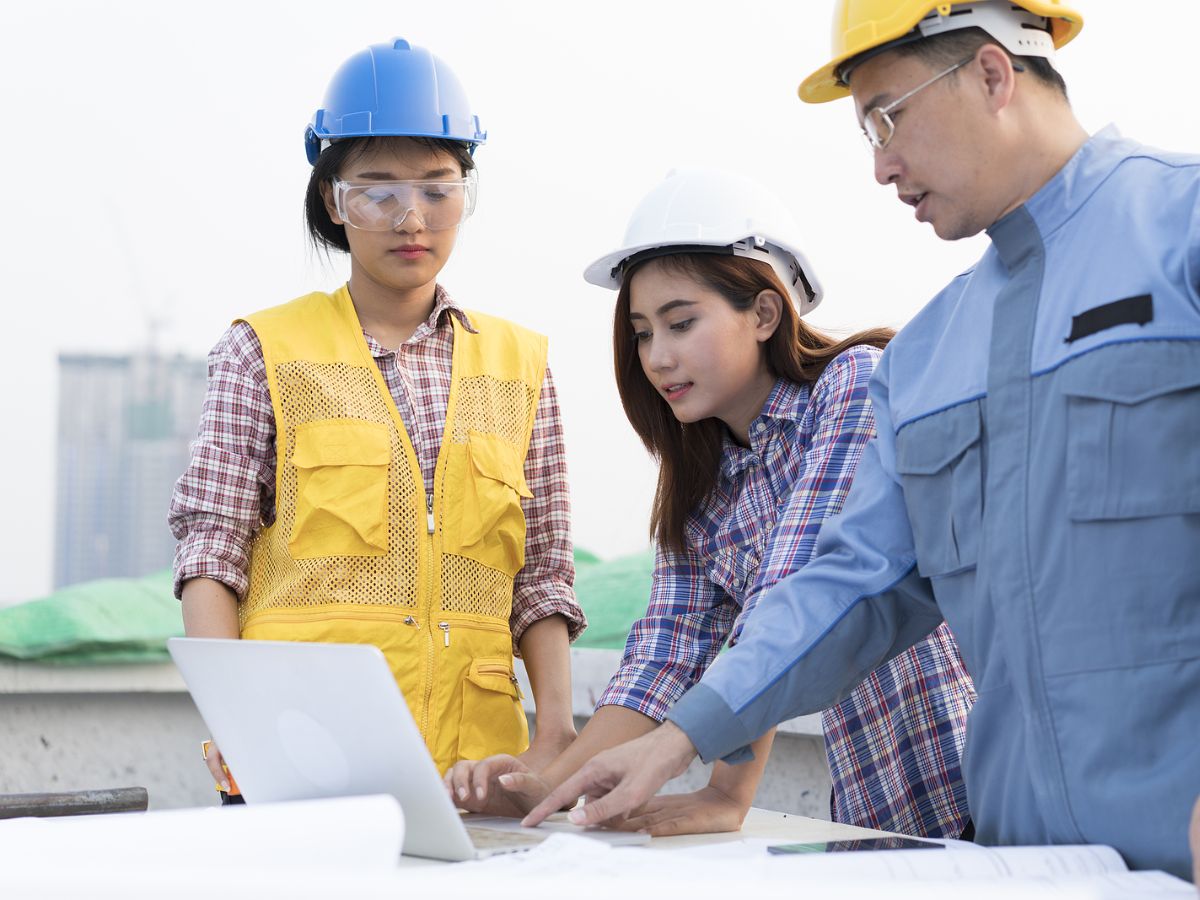 Asian man in hard hat pointing out a laptop while two female women in two hard hats look on