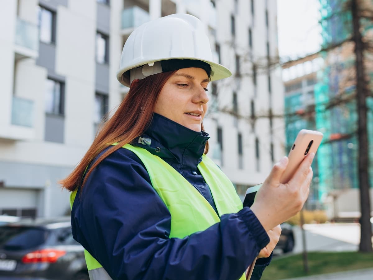 A woman in a hard hat and safety vest holding a cell phone, ready to communicate on the job site.